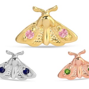 May include: Three moth-shaped jewelry pieces in gold, silver, and rose gold. The gold moth has pink gems, the silver has blue gems, and the rose gold has green gems. The jewelry is likely earrings or pins.