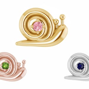 May include: Three snail-shaped jewelry pieces. One is gold with a pink gemstone, another is rose gold with a green gemstone, and the third is silver with a blue gemstone. Each snail has a spiraled shell and two small antennae.