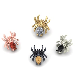 May include: Four spider-shaped jewelry pieces in various metallic finishes: rose gold with a red gem, gold with a blue gem, silver with a black gem, and black with a gold gem. The spiders have detailed legs and bodies.