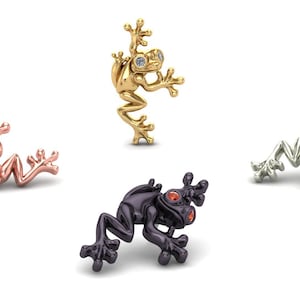 May include: Four frog-shaped pins or ornaments in different metallic finishes: a rose gold frog with a green eye, a yellow gold frog with a clear stone eye, a black metal frog with red eyes, and a silver-toned frog with dark eyes.