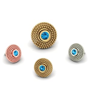 May include: Four pieces of jewelry, each with a central blue gemstone. The pieces are in gold, rose gold, and silver tones, with a beaded texture around the gemstone. The jewelry is set against a white background.