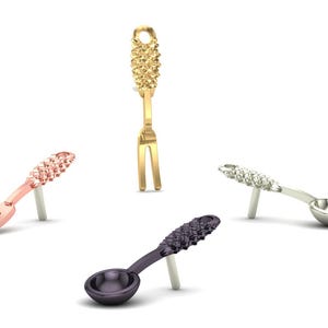 May include: Four miniature decorative utensils: a gold fork, a rose gold fork, a silver spoon, and a dark purple spoon. Each utensil has a textured handle and is propped up on a small stand.