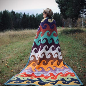 Może przedstawiać: Osoba w kolorowym, dzianym kocu z falistym wzorem. Koc wykonany jest z różnych kolorów, w tym pomarańczowego, żółtego, zielonego, niebieskiego, fioletowego i brązowego. Osoba stoi na polu z drzewami w tle.