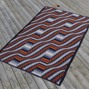 Könnte beinhalten: Eine rechteckige gewebte Decke mit einem geometrischen Muster aus wellenförmigen Linien und rechteckigen Blöcken. Die Decke hat eine Farbpalette aus Orange, Blau, Weiß und Marineblau. Die Decke liegt auf einer Holzterrasse.