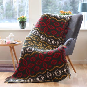 May include: A green, red, white, and gold crocheted throw blanket with fringe on a gray armchair. The blanket has a geometric pattern.