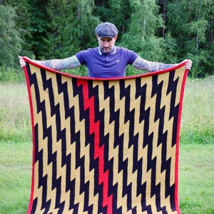Könnte beinhalten: Eine rechteckige Strickdecke mit einem sich wiederholenden schwarzen und beigen Zickzackmuster auf der Oberfläche. Ein einzelner vertikaler roter Zickzackstreifen verläuft durch die Mitte des Designs. Die Decke ist mit einem roten Rand eingefasst.