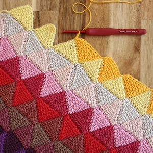 Peut inclure: Une couverture au crochet faite à la main en cours, avec un motif géométrique de triangles aux tons de jaune, rose, rouge et violet. Un crochet rouge et du fil jaune sont visibles sur une surface en bois.