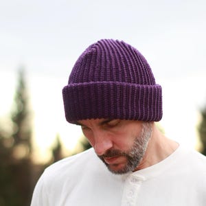 Op de afbeelding: Een close-up van een paarse geribbelde gebreide beanie. De hoed is een diepe tint paars en heeft een omgeslagen rand. De beanie is gemaakt van een dik, getextureerd garen, perfect voor koud weer.