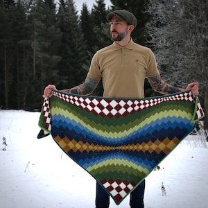 Op de afbeelding: Een kleurrijke gehaakte sjaal met een geometrisch patroon in tinten groen, blauw, bruin en rood. De sjaal wordt vastgehouden door een persoon die een bruin poloshirt en een groene pet draagt.