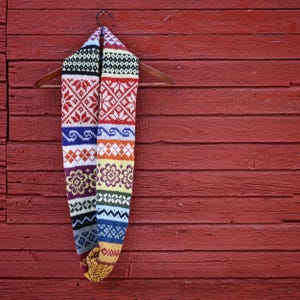 Pode incluir: Um cachecol infinito colorido e estampado pendurado em um cabide de madeira contra um fundo de madeira vermelha. O cachecol apresenta vários desenhos geométricos e florais em vermelho, branco, azul, amarelo e roxo. O cachecol é feito de material de malha.