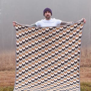 Könnte beinhalten: Eine rechteckige Decke mit einem sich wiederholenden Zickzackmuster in Braun-, Grau- und Cremetönen. Die Decke hat einen grauen Rand. Die Decke wird von einer Person gehalten, die ein weißes T-Shirt und eine lila Mütze trägt. Die Decke ist aus weichem Material.