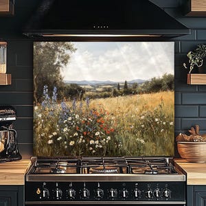 Puede incluir: Un protector de salpicaduras de cocina con una pintura de paisaje. La obra de arte representa un campo de flores silvestres en tonos rojos, blancos y azules, con colinas onduladas y un cielo nublado. La pintura está montada sobre una cocina de gas negra.