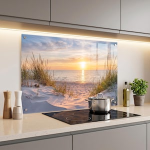 May include: A rectangular glass kitchen backsplash panel featuring a printed image of a sandy beach with tall dune grass at sunset. The scene shows a calm ocean under a golden sky. The panel is mounted behind a black induction cooktop.