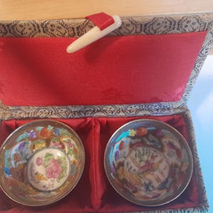 Puede incluir: Dos cuencos pequeños, redondos, de porcelana con diseños florales y de pájaros coloridos dentro de una caja de madera forrada de rojo con un exterior de tela dorada.
