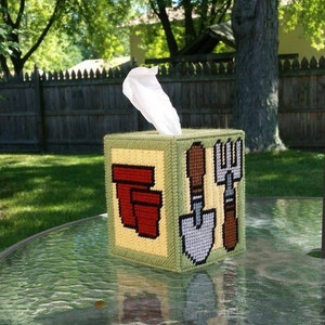 May include: A green and yellow tissue box cover with a red, yellow, and white gardening theme. The cover features a red flower pot, a gray shovel, a brown rake, and a brown trowel.