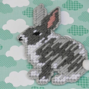 May include: A gray and white yarn bunny with a pink nose and a white pom-pom tail. The bunny is sitting on a green and white patterned background.