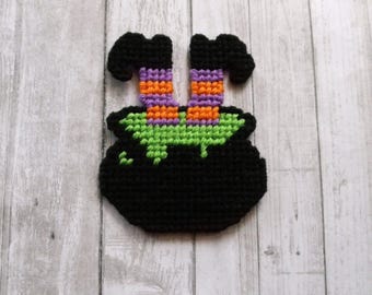 May include: A black crocheted cauldron with a bright green liquid spilling over the rim. Two legs extend upward from the cauldron, featuring horizontal stripes in alternating purple and orange colors, ending in black feet pointing outward.