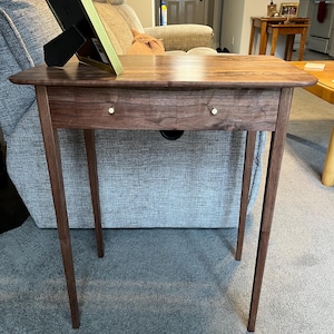 May include: A dark brown wooden end table with two drawers and a simple design. The table has a smooth, polished finish and is supported by four slender legs. The table is sitting on a gray carpet.