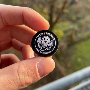 Peut inclure: Épinglette ronde noire et blanche avec un fantôme faisant un doigt d'honneur et le texte "KEIN STANDARD MADE IN THURINGIA".