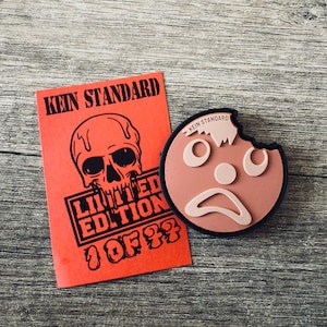 Könnte beinhalten: Ein roter Patch mit Totenkopf und Knochen und dem Text "KEIN STANDARD LIMITED EDITION 1 OF 77". Ein brauner Patch mit einem traurigen Gesicht und dem Text "KEIN STANDARD" befindet sich neben dem roten Patch.