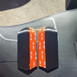 Puede incluir: Dos componentes de plástico rígido negro, angulados, cada uno con una tira vertical de cinta adhesiva naranja 3M VHB aplicada en el centro. Las piezas tienen un acabado mate liso y una forma doblada distintiva para el montaje.