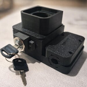 Puede incluir: Un dispositivo negro impreso en 3D con una parte superior cuadrada, una cerradura de llave y dos llaves. El dispositivo tiene una superficie texturizada y parece ser un componente o pieza hecho a medida. Las llaves están unidas a un pequeño anillo de metal.