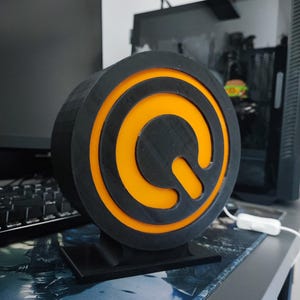 Peut inclure: Un objet décoratif circulaire noir et orange avec un support. L'objet présente un design en couches avec un centre orange vif et un anneau extérieur noir. L'objet est sur un bureau avec un ordinateur et un clavier en arrière-plan.