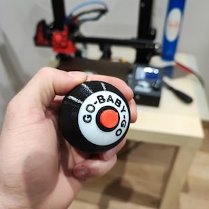 May include: A black and white 3D printed sphere with a red button and the text "GO-BABY-GO" printed on it.