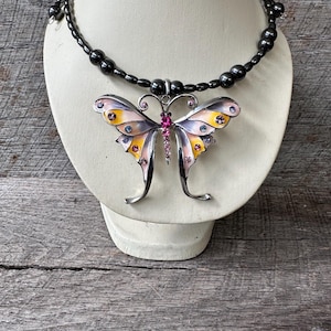 May include: A black beaded necklace with a large butterfly pendant. The butterfly is made of metal and has pink, yellow, and blue enamel accents. The butterfly has clear rhinestones on its wings.