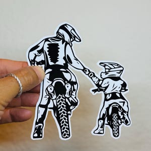 May include: Black and white sticker featuring a parent and child on dirt bikes. The parent is holding the child's hand, both wearing helmets. The sticker has a white background and is ideal for motocross enthusiasts.