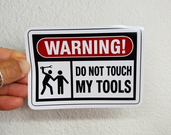 May include: A rectangular sticker with a black border and the words "WARNING! DO NOT TOUCH MY TOOLS" in white text. The top section has a red oval with the word "WARNING!" in white. Below is a graphic of two stick figures.