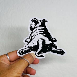 May include: A black and white sticker of a bulldog, viewed from behind. The sticker depicts the dog's back, legs, and tail in a stylized illustration. The sticker is held by a hand with silver rings.