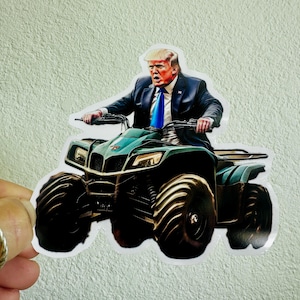 Puede incluir: Una pegatina que muestra a una persona con traje y corbata azul montando un quad verde azulado. La persona tiene el pelo rubio y mira hacia adelante con la boca abierta. El quad tiene neumáticos negros grandes.