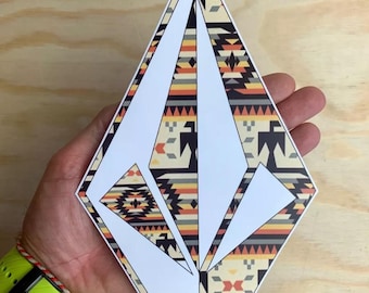 Könnte beinhalten: Ein weißer, rautenförmiger Aufkleber mit einem geometrischen Muster in Schwarz, Orange, Gelb und Grau. Der Aufkleber befindet sich auf einer Holzoberfläche.