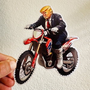 Op de afbeelding: Een sticker van een man met blond haar die op een rode en witte crossmotor rijdt. De man draagt een donker pak en een rode stropdas. De motor heeft een Amerikaans vlagontwerp aan de zijkant.