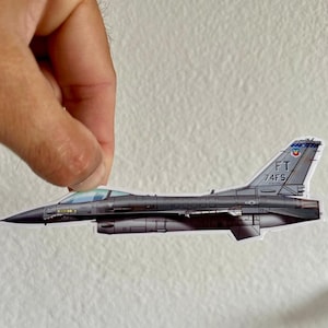 May include: A gray and white F-16 fighter jet with the text "FT 74FS" on the tail. The jet is a sticker with a slightly faded appearance.