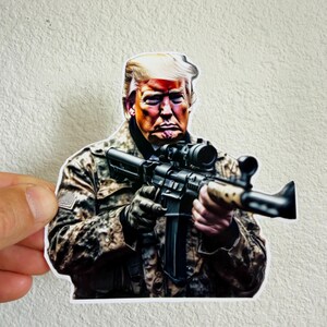 May include: A sticker featuring a digitally illustrated image of a person holding a rifle. The person is wearing a camouflage jacket. The sticker is held by a hand.