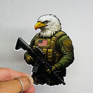 May include: A sticker featuring an eagle with a white head and yellow beak, wearing a green military uniform and holding a black rifle. The uniform has a US flag patch. The sticker is held by a hand.