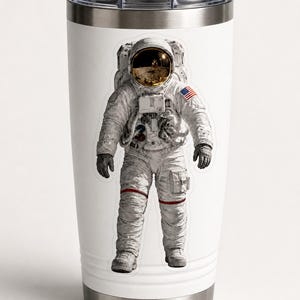 Op de afbeelding: Witte reismok met een afbeelding van een astronaut in een ruimtepak. De astronaut draagt een wit ruimtepak met een goud getinte helm en de Amerikaanse vlag op de arm. De mok heeft een zilveren basis en deksel.