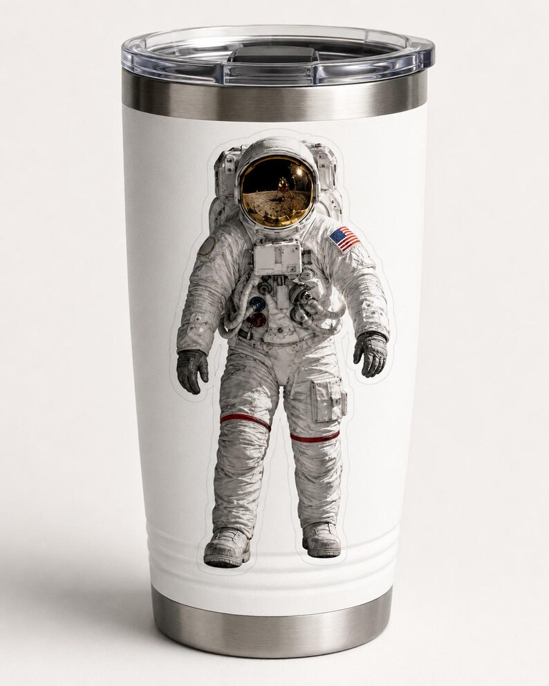 Op de afbeelding: Witte reismok met een afbeelding van een astronaut in een ruimtepak. De astronaut draagt een wit ruimtepak met een goud getinte helm en de Amerikaanse vlag op de arm. De mok heeft een zilveren basis en deksel.
