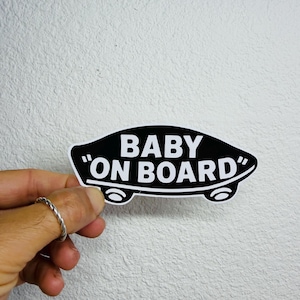 Könnte beinhalten: Ein schwarz-weißer "Baby On Board" Aufkleber. Der Aufkleber hat die Form eines Autos mit dem Text in einer fetten, serifenlosen Schriftart. Der Aufkleber wird vor einer weißen, strukturierten Wand gehalten.