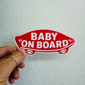 Könnte beinhalten: Ein rot-weißer "Baby On Board" Aufkleber. Der Aufkleber hat die Form eines Autos und trägt die Worte "BABY ON BOARD" in weißen Großbuchstaben. Der Aufkleber wird von einer Hand gehalten.