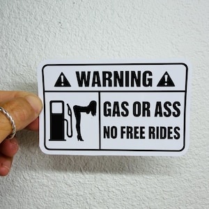 以下が含まれることがあります： 黒いテキストとグラフィックが施された長方形の白いステッカー。ステッカーには上部に「WARNING」と書かれており、ガソリンスタンドと女性が描かれています。下のテキストは「GAS OR ASS NO FREE RIDES」と書かれています。