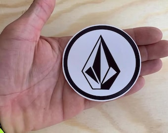 Könnte beinhalten: Ein weißer runder Aufkleber mit schwarzem Rand und einem schwarzen rautenförmigen Logo in der Mitte.