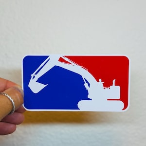 May include: A rectangular sticker with a blue and red background. A white excavator silhouette is in the center. The sticker is held by a hand with a silver ring.