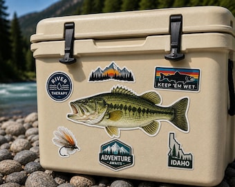 Puede incluir: Una nevera portátil beige adornada con varias pegatinas temáticas de actividades al aire libre, incluyendo un pez grande, montañas y el texto "Keep 'em Wet". La nevera está situada cerca de un río y rocas, sugiriendo pesca o camping.