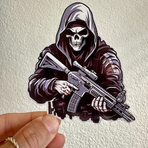 May include: A sticker featuring a hooded figure with a skull face holding a rifle. The figure is wearing a black tactical vest and has a scope on the rifle.