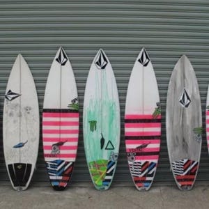 K&ouml;nnte beinhalten: Sieben Surfboards mit verschiedenen bunten Designs, darunter rosa, gr&uuml;n und schwarz. Die Surfboards sind an einer grauen Metallwand aufgereiht.