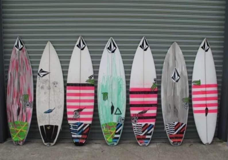 K&ouml;nnte beinhalten: Sieben Surfboards mit verschiedenen bunten Designs, darunter rosa, gr&uuml;n und schwarz. Die Surfboards sind an einer grauen Metallwand aufgereiht.