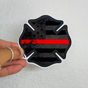 May include: A black and gray sticker in the shape of a Maltese cross, featuring a distressed American flag design with a red stripe. The sticker has a white border and is held by a hand.